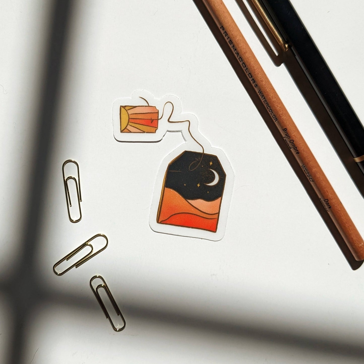 A vinyl sticker in the shape of a teabag with a desert landscape in the bag and a sunrise on the tag. The sticker is on a white background with a pen, pencil, paperclips, and artistic shadows.